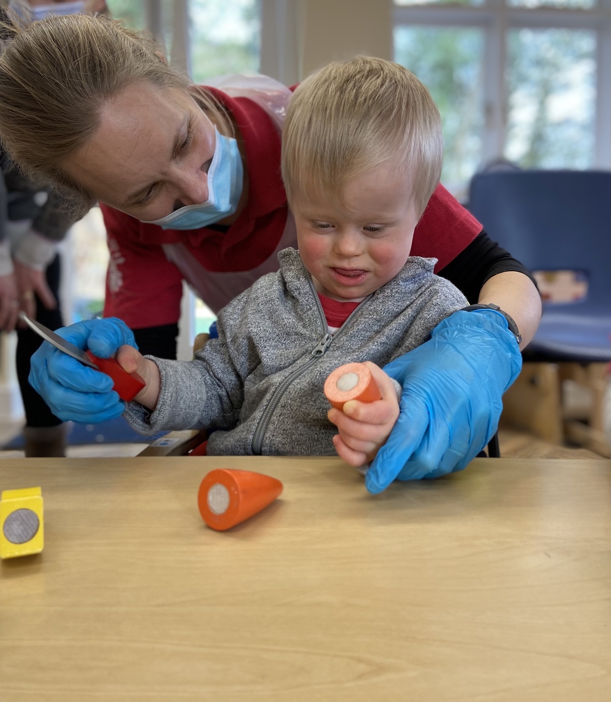 A little boy is using a play cooking set, slicing  wooden carrots, with help from a Playskill therapist